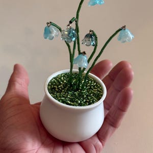 May include: A miniature beaded lily of the valley plant in a white pot. The flowers are light blue and the stems are green. The pot is filled with green beads. The plant is held in a hand.