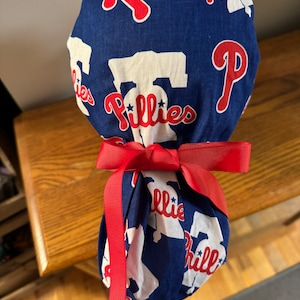 May include: A blue and white fabric scrub cap with a red ribbon tied around it. The fabric has a repeating pattern of the Philadelphia Phillies logo, which includes the letter "P" and the word "Phillies".