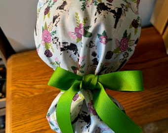 May include: A white scrub cap with a floral and cow print design. The cap is tied with a green ribbon.