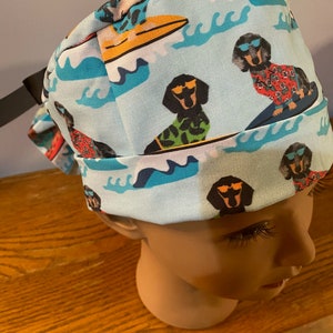 May include: Light blue surgical scrub cap featuring a repeating pattern of dachshunds wearing sunglasses and Hawaiian shirts, surfing on colorful surfboards. The design includes white waves and surfboards in red and yellow.