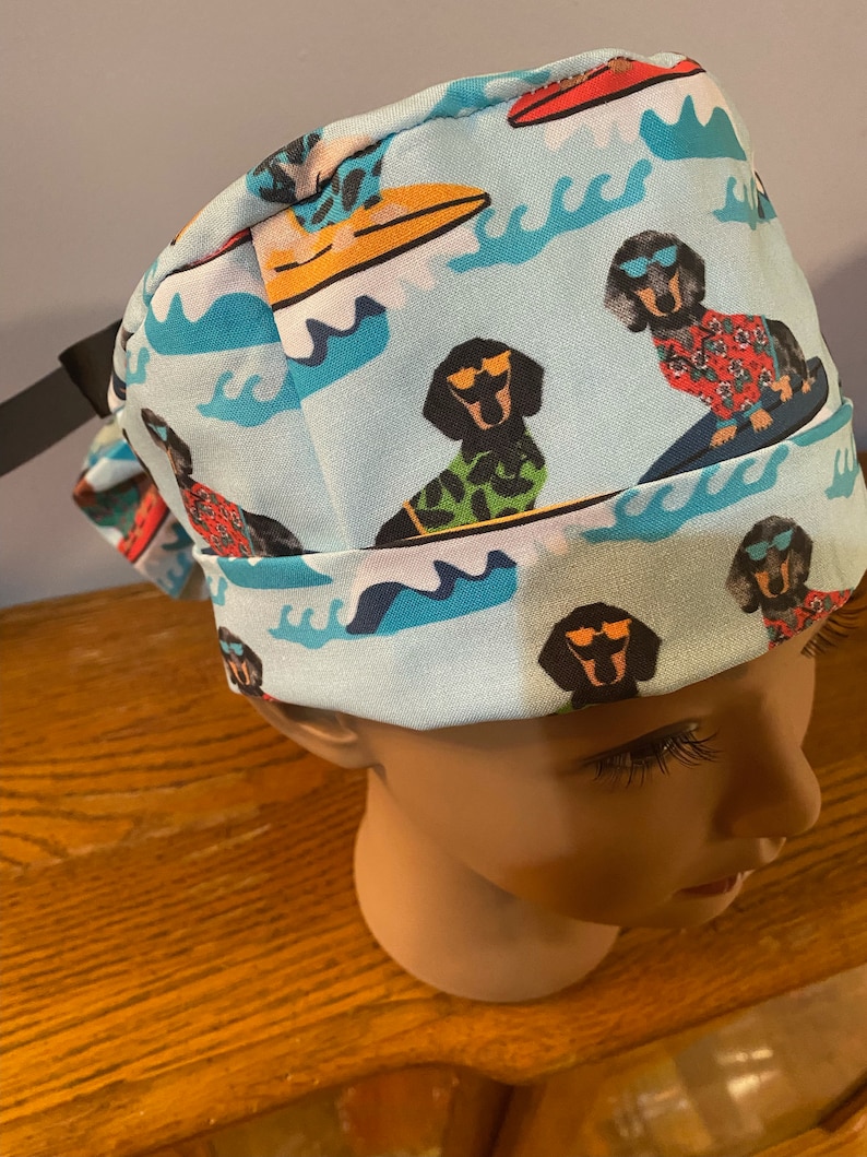 May include: Light blue surgical scrub cap featuring a repeating pattern of dachshunds wearing sunglasses and Hawaiian shirts, surfing on colorful surfboards. The design includes white waves and surfboards in red and yellow.