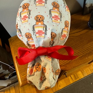 May include: A light blue fabric scrub hat with a repeating pattern of brown dogs sitting in red and yellow "Cup Noodles" containers. The hat is tied with a red ribbon.