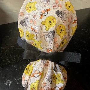 May include: A beige scrub cap featuring a pattern of yellow Winnie the Pooh, Eeyore, and Piglet faces. The cap is tied with a large black bow.