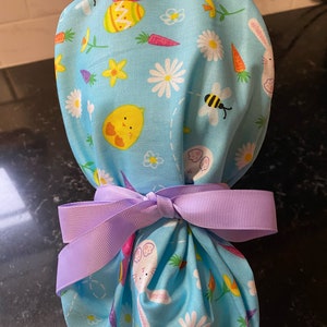 May include: A blue fabric Easter-themed scrub hat with a purple ribbon tied around it. The hat has a pattern of bunnies, chicks, carrots, and eggs.
