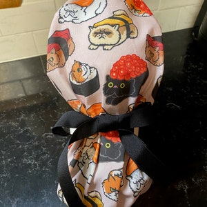 May include: A pink fabric scrub hat with a black ribbon tied around it. The hat has a pattern of cats dressed as sushi rolls.