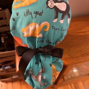 May include: A turquoise blue fabric shower cap with a black ribbon tied around it. The fabric has a repeating pattern of cats in various poses with the text "kitty squat", "Purr-fect", and "Tail Be" visible.