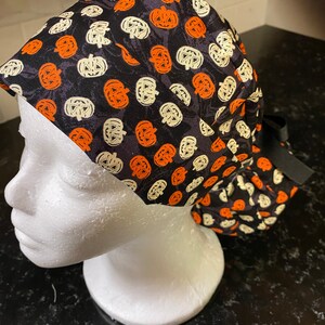 Puede incluir: Un gorro de quir&oacute;fano negro con un patr&oacute;n de calabazas naranjas y blancas. El gorro tiene un lazo negro.