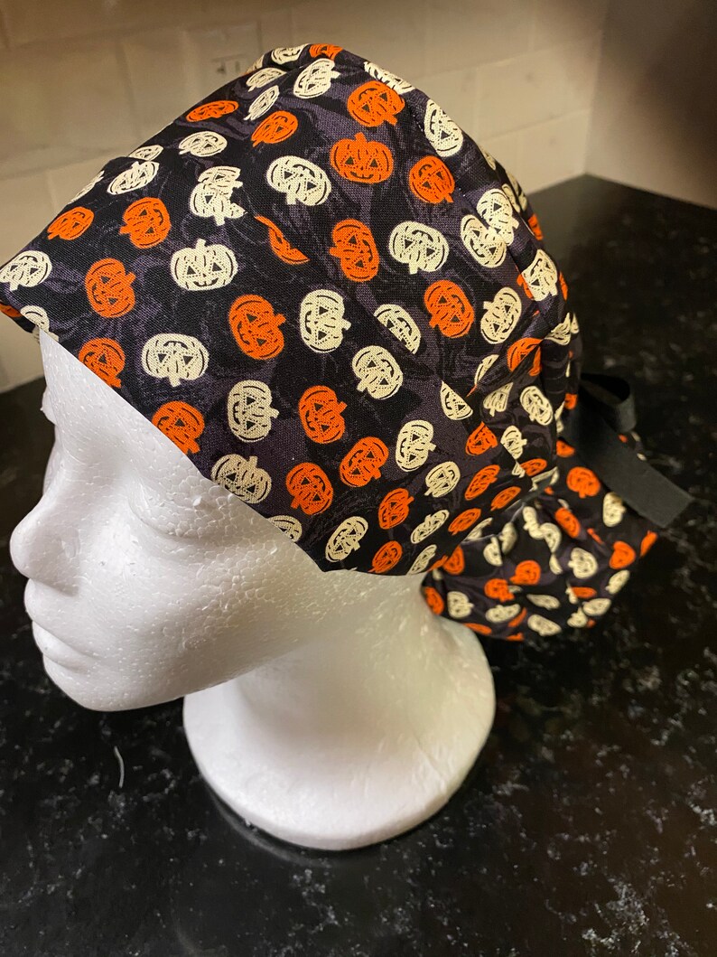 Puede incluir: Un gorro de quir&oacute;fano negro con un patr&oacute;n de calabazas naranjas y blancas. El gorro tiene un lazo negro.
