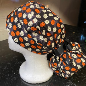 Puede incluir: Un gorro de quir&oacute;fano negro con un patr&oacute;n de calabaza blanca y naranja. El gorro tiene una cinta negra atada en la parte posterior.