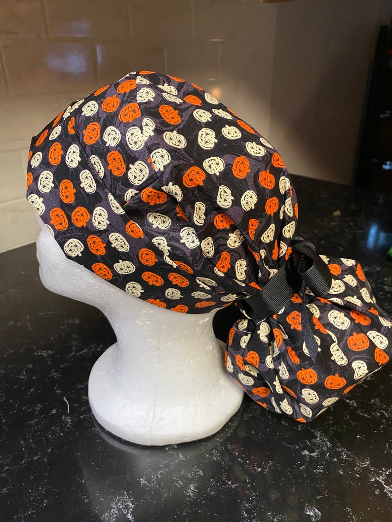 Puede incluir: Un gorro de quir&oacute;fano negro con un patr&oacute;n de calabaza blanca y naranja. El gorro tiene una cinta negra atada en la parte posterior.