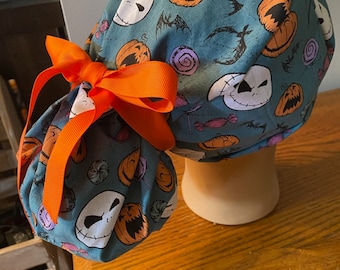 Könnte beinhalten: Eine tealblaue OP-Mütze mit orangefarbenem Schleifenband. Die Mütze hat ein Halloween-Thema mit einem sich wiederholenden Muster aus Jack Skellington-Gesicht, Kürbissen und Süßigkeiten.