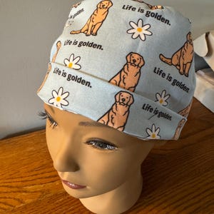 May include: A light blue scrub hat with a repeating pattern of golden retrievers and daisies. The text "Life is golden." is repeated throughout the pattern.