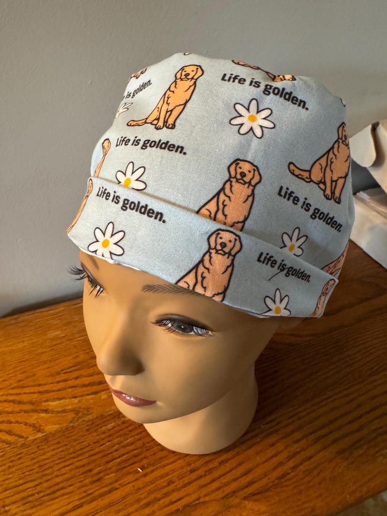May include: A light blue scrub hat with a repeating pattern of golden retrievers and daisies. The text "Life is golden." is repeated throughout the pattern.