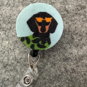 May include: A round, light blue fabric badge reel featuring a black dachshund wearing orange sunglasses. The dog is wearing a green and black patterned shirt. The badge reel has a clear plastic clip and a metal retractable cord.