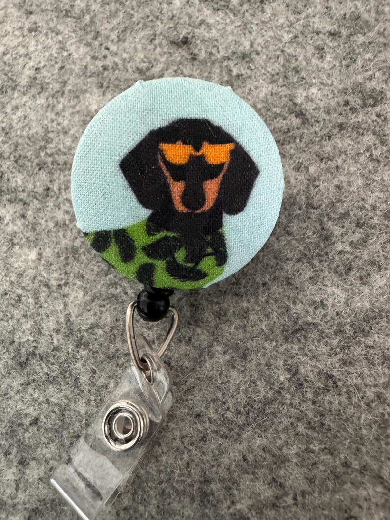 May include: A round, light blue fabric badge reel featuring a black dachshund wearing orange sunglasses. The dog is wearing a green and black patterned shirt. The badge reel has a clear plastic clip and a metal retractable cord.