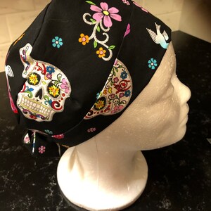 May include: Black scrub hat with a colourful sugar skull design, pink flowers, and a white bird. The hat has a tie at the back.