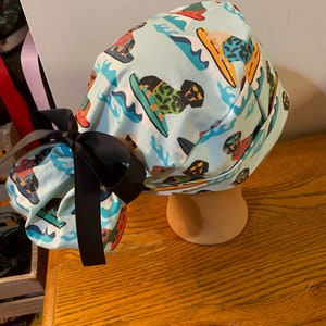May include: A light blue surgical scrub cap featuring a repeating pattern of dogs surfing on colorful surfboards. The cap is tied with a black bow. The cap is displayed on a mannequin head.