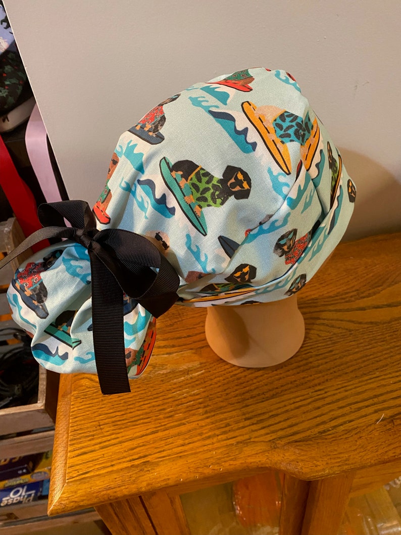 May include: A light blue surgical scrub cap featuring a repeating pattern of dogs surfing on colorful surfboards. The cap is tied with a black bow. The cap is displayed on a mannequin head.