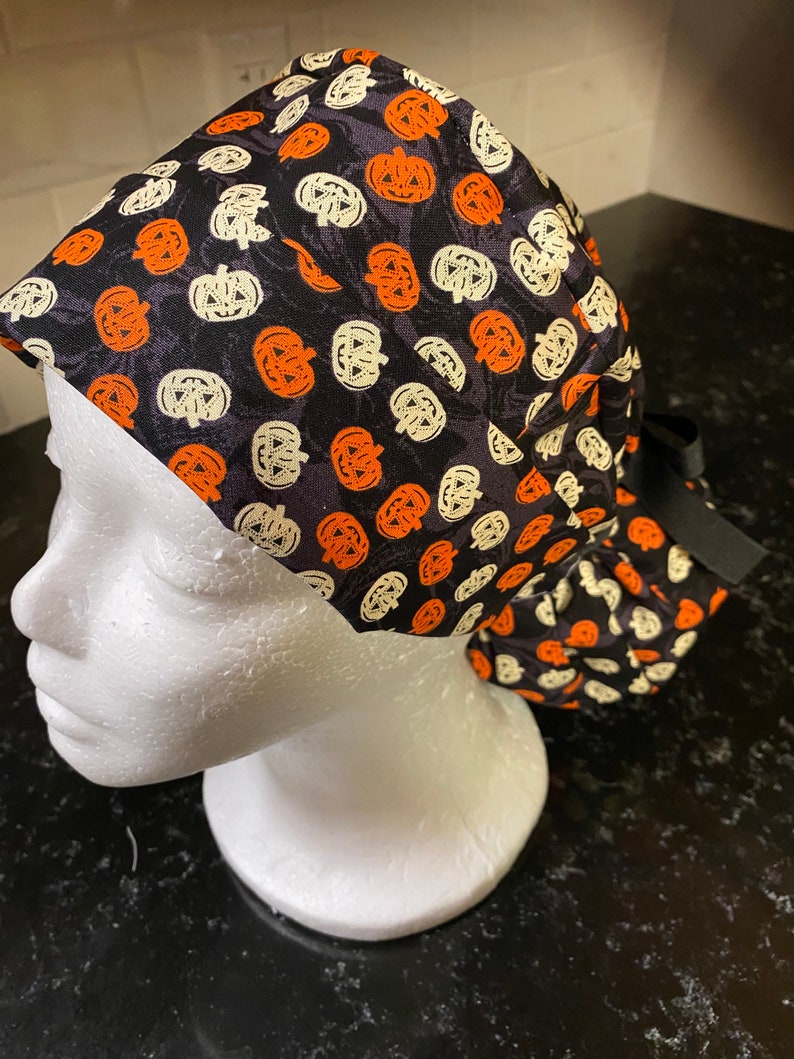 Puede incluir: Un gorro de quir&oacute;fano negro con un patr&oacute;n de calabazas naranjas y blancas. El gorro tiene una cinta negra.