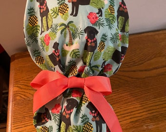 May include: A blue and green patterned surgical scrub hat with black dogs wearing leis and holding pineapples. The hat has a red bow tied around it.