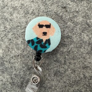 May include: A round badge reel featuring a cartoon dog wearing sunglasses and a patterned shirt. The badge reel has a light blue background and a clear plastic clip. The dog's face is tan, and the shirt is teal and black.