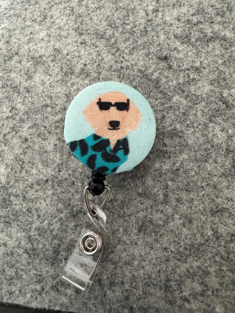 May include: A round badge reel featuring a cartoon dog wearing sunglasses and a patterned shirt. The badge reel has a light blue background and a clear plastic clip. The dog's face is tan, and the shirt is teal and black.