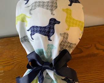 May include: A white fabric scrub cap with a repeating pattern of dachshund dogs in navy, grey, and yellow-green. The cap is tied with a navy blue ribbon. The cap is designed for medical professionals.