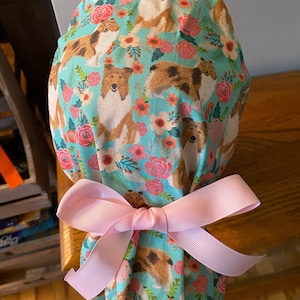 May include: A teal scrub cap featuring a repeating pattern of brown and white dogs and pink flowers. The cap is tied with a light pink bow. The fabric has a floral and dog theme.