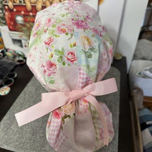 May include: A floral patterned scrub cap featuring a patchwork design with pink roses and green leaves on a white background. The cap is tied with a light pink ribbon bow. The cap is designed for medical professionals.