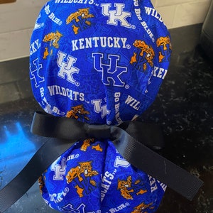 May include: A blue and white fabric scrub hat with a black ribbon tied around it. The fabric has a pattern of the University of Kentucky Wildcats logo, the words "Go Big Blue", and "Wildcats".