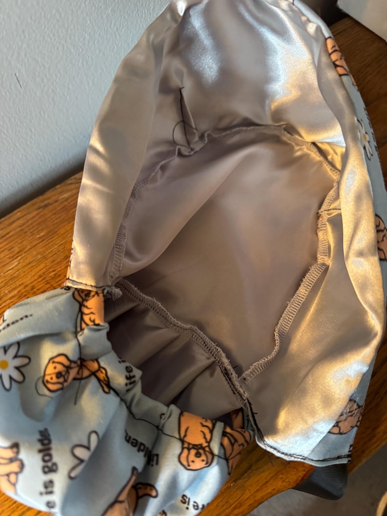 May include: A light blue satin scrub hat with a print of golden retrievers and the text "Life is golden" and "Life is unique". The hat has a gray satin lining.