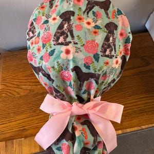May include: A teal scrub cap featuring a repeating pattern of brown and white dogs and pink and orange flowers. The cap is tied with a light pink bow.