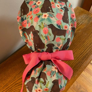 May include: A turquoise blue fabric scrub hat with a floral and chocolate lab print. The hat is tied with a pink ribbon.
