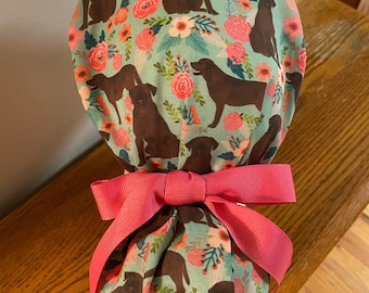 May include: A turquoise blue fabric scrub hat with a floral and chocolate lab print. The hat is tied with a pink ribbon.