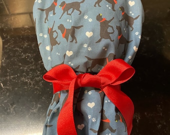 May include: A blue fabric scrub hat with a pattern of black labrador retrievers wearing red bandanas and white hearts. The hat is tied with a red ribbon.