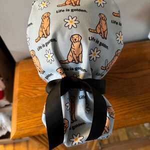 May include: A blue fabric pouch with a black ribbon tie. The pouch is decorated with a repeating pattern of golden retrievers and daisies. The text "Life is golden." is repeated throughout the pattern.