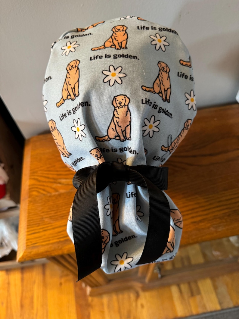 May include: A blue fabric pouch with a black ribbon tie. The pouch is decorated with a repeating pattern of golden retrievers and daisies. The text "Life is golden." is repeated throughout the pattern.