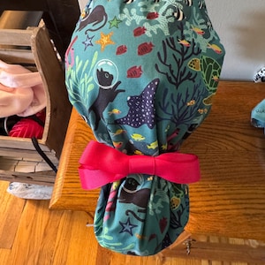 May include: A teal scrub cap featuring an underwater scene with black cats in space helmets, fish, coral, and a stingray. The cap is tied with a bright pink bow.