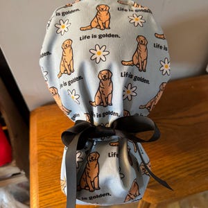 May include: A blue fabric bag with a drawstring closure and a black ribbon tied around it. The bag is decorated with a pattern of golden retrievers and daisies. The text "Life is golden." is repeated throughout the pattern.