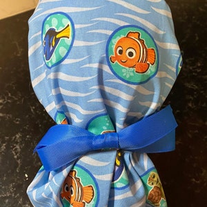 May include: Blue and white scrub hat with a Finding Nemo theme. The hat features a blue background with white waves and circular designs featuring Dory, Nemo, and a turtle. The hat is tied with a blue ribbon.