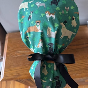 Könnte beinhalten: Grüne OP-Haube mit einem Muster aus verschiedenen Hunderassen und Kleeblättern. Die Haube ist mit einer schwarzen Schleife gebunden. Der Stoff zeigt Illustrationen von Hunden in verschiedenen Posen und Farben.