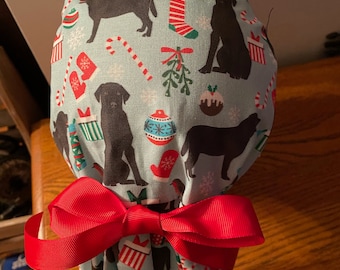 May include: A blue and white patterned surgical scrub hat with a red bow. The pattern features black dogs, candy canes, ornaments, and other Christmas-themed designs.