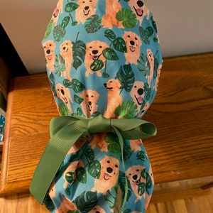May include: A teal scrub cap featuring a repeating pattern of smiling golden retrievers and green monstera leaves. The cap is tied with a green ribbon bow. The cap is designed for medical professionals.
