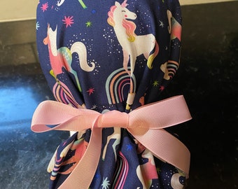 May include: A blue scrub hat with a unicorn print and a pink ribbon tied around it. The unicorns are white with pink, yellow, and blue accents. The background is a dark blue with white stars.