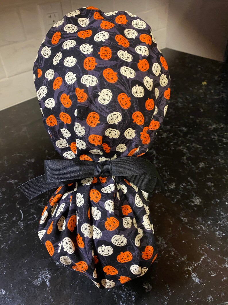 Puede incluir: Un gorro de quir&oacute;fano negro con un patr&oacute;n de calabazas blancas y naranjas. El gorro est&aacute; atado con una cinta negra.