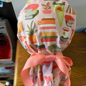 May include: Pink scrub hat with a print of books, plants, and glasses. The hat has a pink ribbon tied around it and the text "I ♥ Books" on the front.