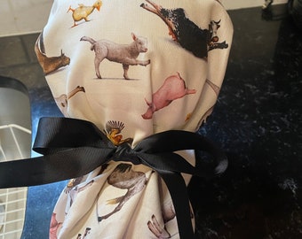 May include: A white fabric scrub cap with a black ribbon tied around it. The cap features a pattern of various farm animals, including pigs, cows, horses, and chickens.