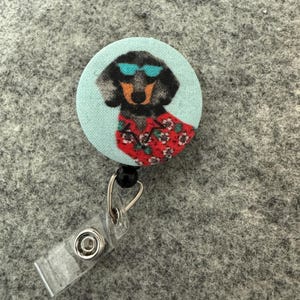 May include: A round, light blue retractable badge reel featuring a dachshund wearing sunglasses and a Hawaiian shirt. The dog is black and brown with a red floral shirt. The reel has a clear plastic clip and a black bead.