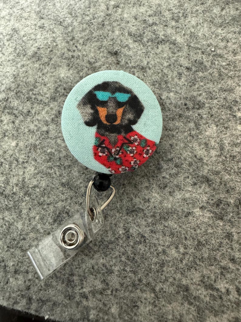 May include: A round, light blue retractable badge reel featuring a dachshund wearing sunglasses and a Hawaiian shirt. The dog is black and brown with a red floral shirt. The reel has a clear plastic clip and a black bead.