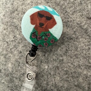 May include: A round retractable badge reel featuring a cartoon dachshund wearing sunglasses and a Hawaiian shirt. The dog is brown with a light blue background and a black retractable cord and clip.